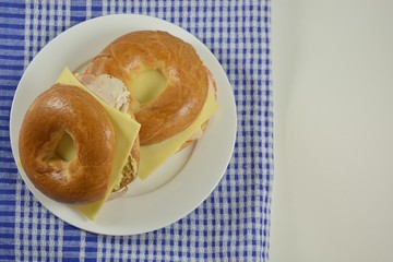 Cheese and chicken with egg salad on a bagel on a white plate on a checked cloth in front of a white background