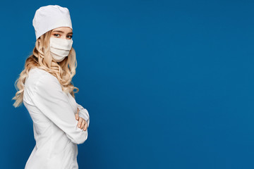 Young female doctor in white coat and medical mask posing by a side, isolated at a blue background with copy space. Nurse in white medical uniform and protective mask posing on blue background