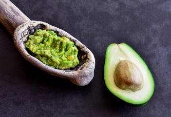 Avocado appetizer in a large wooden spoon and half fruit with a bone, black, vintage background. Avocado Sauce. Gualamole
