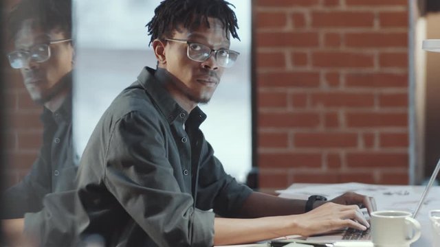 Side view of young African American man in glasses and casual clothes using laptop and the looking at camera and smiling while working at desk in loft office with glass wall