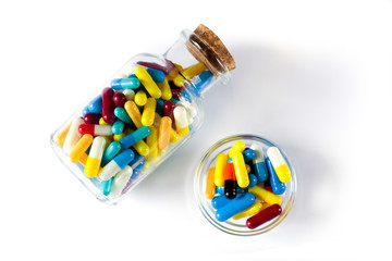 heap of multi-colored medical capsules in a transparent bottle and a small glass bowl on a white background.