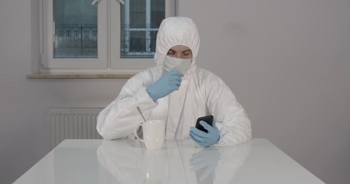 Portrait of young male person stitting behind white dining desk in kitchen in protective suit drinking a cup of tea and reading news on his phone during pandemic coronavirus quarantine time 4K