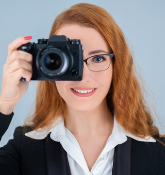 Young redhead woman holding a camera. She is wearing glasses and a suit. 