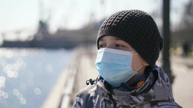 Sportive boy in sanitary mask and jacket standing on a shiny riverbank in spring