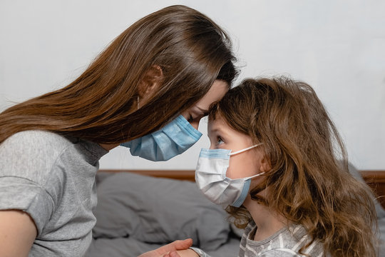 Self isolation, home quarantine. Cute little girl and mother wearing face mask, sitting on bed at home, consoling sad preschool daughter. Concept of coronavirus or COVID-19 pandemic disease symptoms.