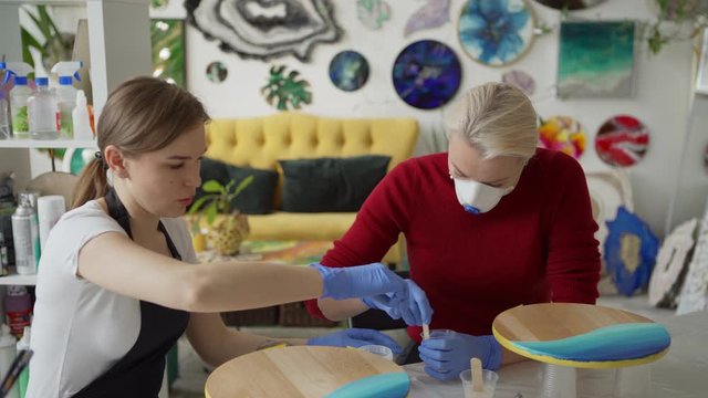 Medium shot of art teacher helping middle aged female student in protective mask to mix epoxy resin for fluid painting sitting at desk in art studio