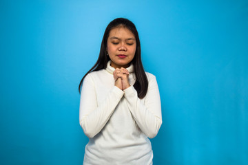 Portrait of Young beautiful asian women using white T-shirt with blue isolated background, folded hand together and pray for covid-19, coronavirus