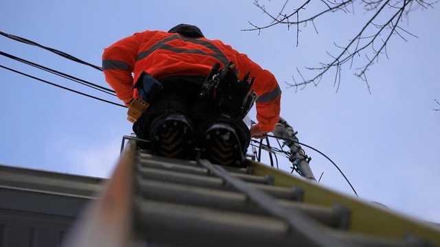 A telecoms operative is seen working from a ladder on a utility pole, wearing high visibility personal protective clothing, high viz PPE, and hard hat