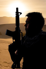 Silhouette of a male soldier resting in the shade on a desert and holding a rifle.  Depicts the...