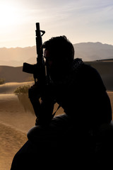 Silhouette of a male soldier resting in the shade on a desert and holding a rifle.  Depicts the...
