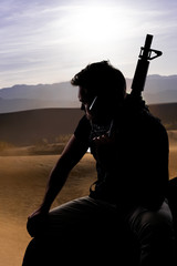 Silhouette of a male soldier resting in the shade on a desert and holding a rifle.  Depicts the...