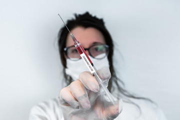 Selective focus shot of syringe held by gloved hand diagonally in center frame filled with red fluid. Background out of focus nurse wearing glasses