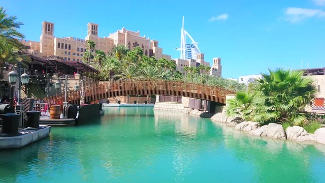 DUBAI, UAE - MARCH 4, 2020: Park area of Souk Madinat Jumeirah market complex with canal, pedestrian bridge, lush greenery, Fort Island, cafes and Burj Al Arab on the background, on March 4 in Dubai