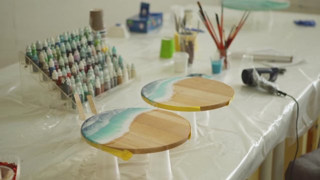 Tracking from above shot of desk with equipment in art studio and two unfinished fluid sea paintings made with epoxy resin and acrylic paints on round wooden canvases