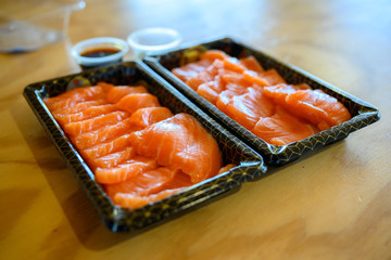Salmon sashimi on a black plastic tray The side has soy sauce and wasabi. Is a delivery set meal On an orange wooden table top.