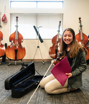 Portrait of smiling teenage girl with violin and sheet music kneeling in classroom