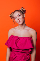 The girl in a red dress on a orange background in the studio. Blonde girl with two hair knots looking to the camera