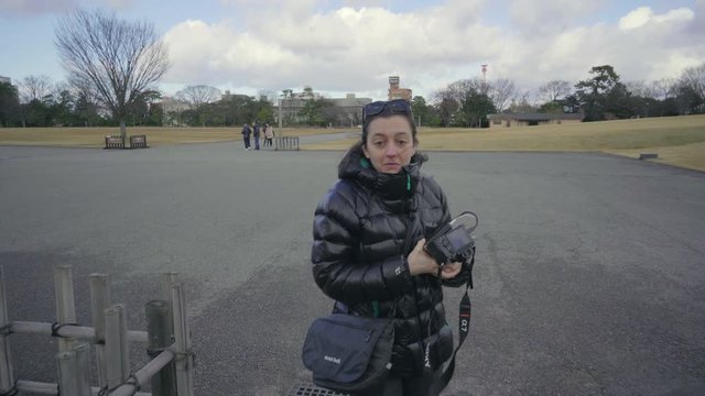 A female, brunette tourist in winter clothing talks to the camera while passing signs at the entrance to Kenrokugen garden via Kanazawa Castle, Japan.