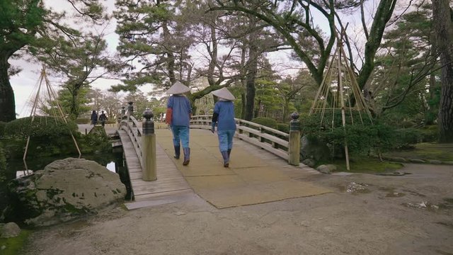 A pair gardeners in uniforms including straw hats cross a bridge over a river in Kenrokuen garden, Kanazawa, Japan.