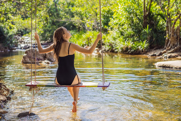 Beautiful happy girl, young pretty cheerful woman traveler sitting, swinging on a swing tied to a tree above the water, smiling in sunny day. Summer vacation, travel concept