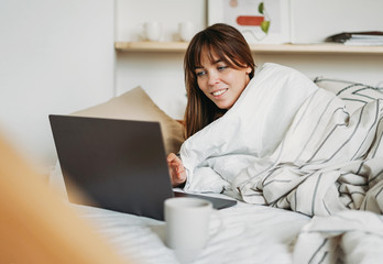 Working young brunette woman freelancer in casual clothes with laptop and cup of tea wake up in bed at home in bright interior stay at home