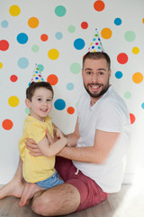 Toddler boy with his father in the birthday hats on a background of wall with colorful circles