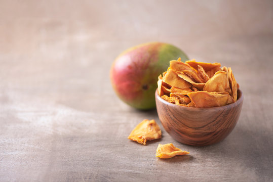 Dried mango in wooden bowl and fresh mango fruit on wood textured background. Copy space. Superfood, vegan, vegetarian food concept. Macro of orange dried mango slices, selective focus. Healthy snack.