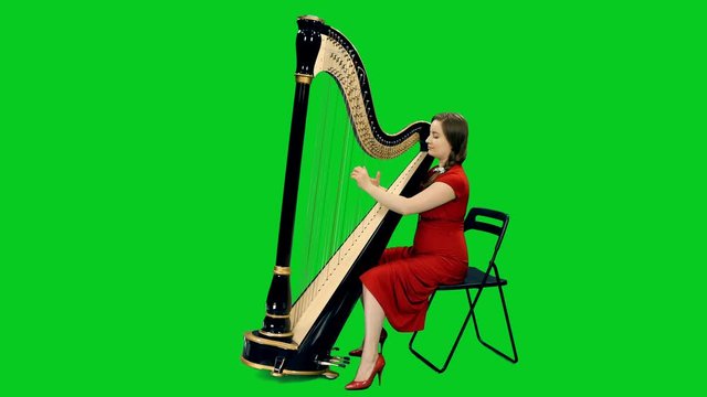 Young female musician in red dress playing harp against green screen background