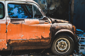 An old orange car that has been left rotting, flat and rusted, parked near a pile of brick blocks.