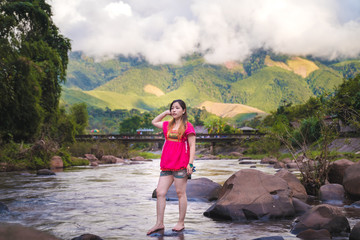 Naklejka premium Beautiful women wearing pink outfits standing on rocks in a stream surrounded by nature, with a background of mountains and clouds.