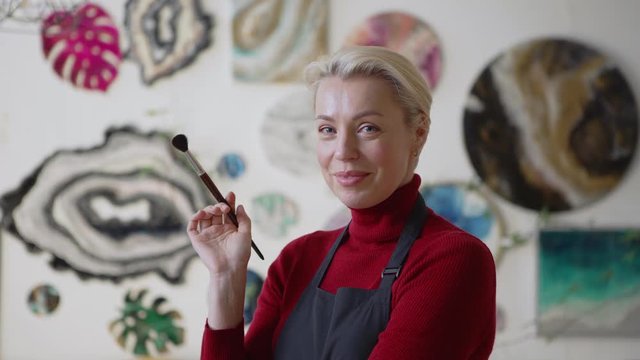 Tilt up side view of beautiful middle aged female artist with blonde short hair thinking standing in art studio with paintbrush in her hand then looking at camera and smiling