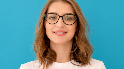 Smart young lady looking at camera and putting on glasses against blue background