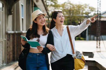 Two happy tourists female friends travel in summer vacations with guide book. group of cheerful sisters walking under sunset time with sun flare. lady point finger showing view and smiling joyful.