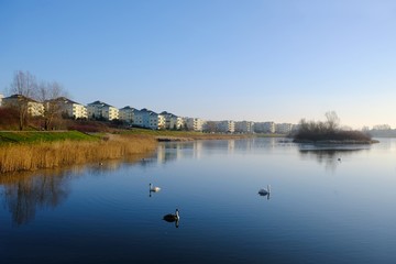 An estate of identical apartment blocks on the lake and its reflection in water, on the outskirts of the city. Idyllic view with blue sky and blue water. Gdansk South, Zakoniczyn, Poland