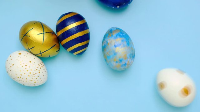 Easter eggs are rolling, knocking each other on blue table. Eggs trendy colored classic blue, white and golden . Happy Easter. Minimal style. Top view.