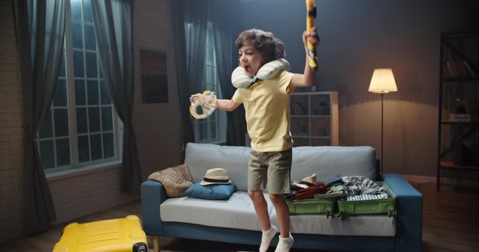 Funny little asian kid with curly hair is jumping on a suitcase tightly packed with clothes, holding mask for scuba diving, excited about upcoming adventure - travelling concept 4k footage