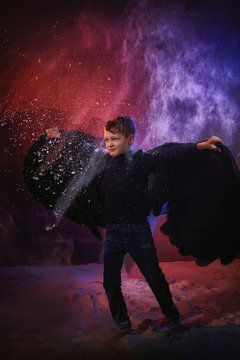 Black Evil Angel On A Dark Background With Colored Lighting. The Concept Of War Between Good And Evil. Boy With Angel Wings During A Photo Shoot With Flour And Loose Powder