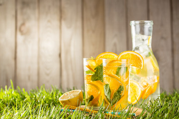 cold lemonade drink with orange and mint in tall glass on green grass lawn wooden background