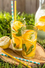cold lemonade drink with orange and mint in tall glass on green grass lawn wooden background