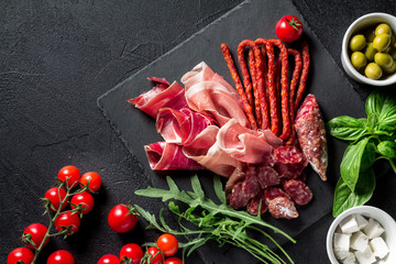 Tapas concept. Prosciutto, sausage chorizo, olives, cherry tomato, arugula, basil and cheese on a black slate board. Antipasti concept.