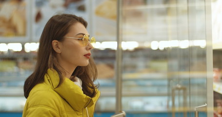 Young Caucasian pretty woman in glasses opening refrigerator and looking at product, then putting...