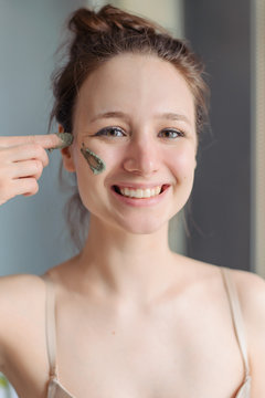 Girl Applies A Green Mask Of Clay, Minerals And Salts To Her Face. A Mask Swab On One Cheek And A Woman 's Finger. Home Light Bathroom. Natural Skin Care. Vegetable Vegan Cosmetics.