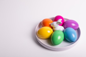 Six Easter eggs in yellow, orange, pink, violet, green, blue colors in the white porcelain plate with white Easter bunny on white background. Copy space, selective focus. Happy Easter!