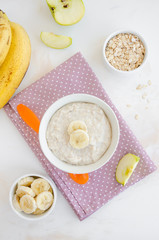 Baby food. Creamy oatmeal with banana slices and apple in a bowl with a spoon on a light background. Healthy breakfast. Porridge for breakfast. Vertical orientation. Top view.
