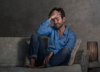 dark and dramatic lifestyle portrait of attractive depressed and worried man at home couch during covid-19 virus outbreak home lockdown feeling sad and overwhelmed