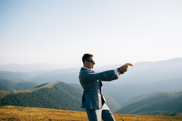 Secret agent in stylish suit standing in mountains,shooting with banana.