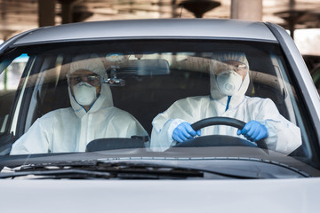 Two men in a car, both wearing virus protective suits