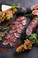 Snacks. Sliced sausage with grapes, figs, Dor blue cheese, arugula, mint, spices and bread on a black background. Background image, copy space