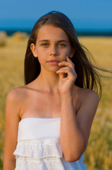 vertical close-up portrait of a cute long-haired green-eyed teenager girl in a white sundress against a blue sky and a wheat field