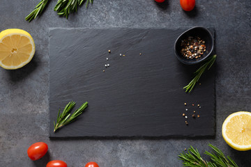 Dark food background with empty black stone board, cherry tomatoes, rosemary, lemon and spicies. Free space for text or product. Top view, flat lay